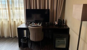 Hotellihuone, jossa on työpöytä, tuoli, televisio ja pieni jääkaappi.