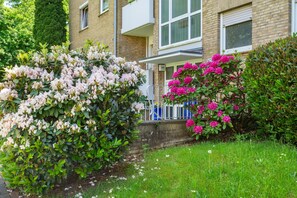 Un edificio residencial con fachada de ladrillo, un balcón y un jardín bien cuidado con arbustos florecientes.