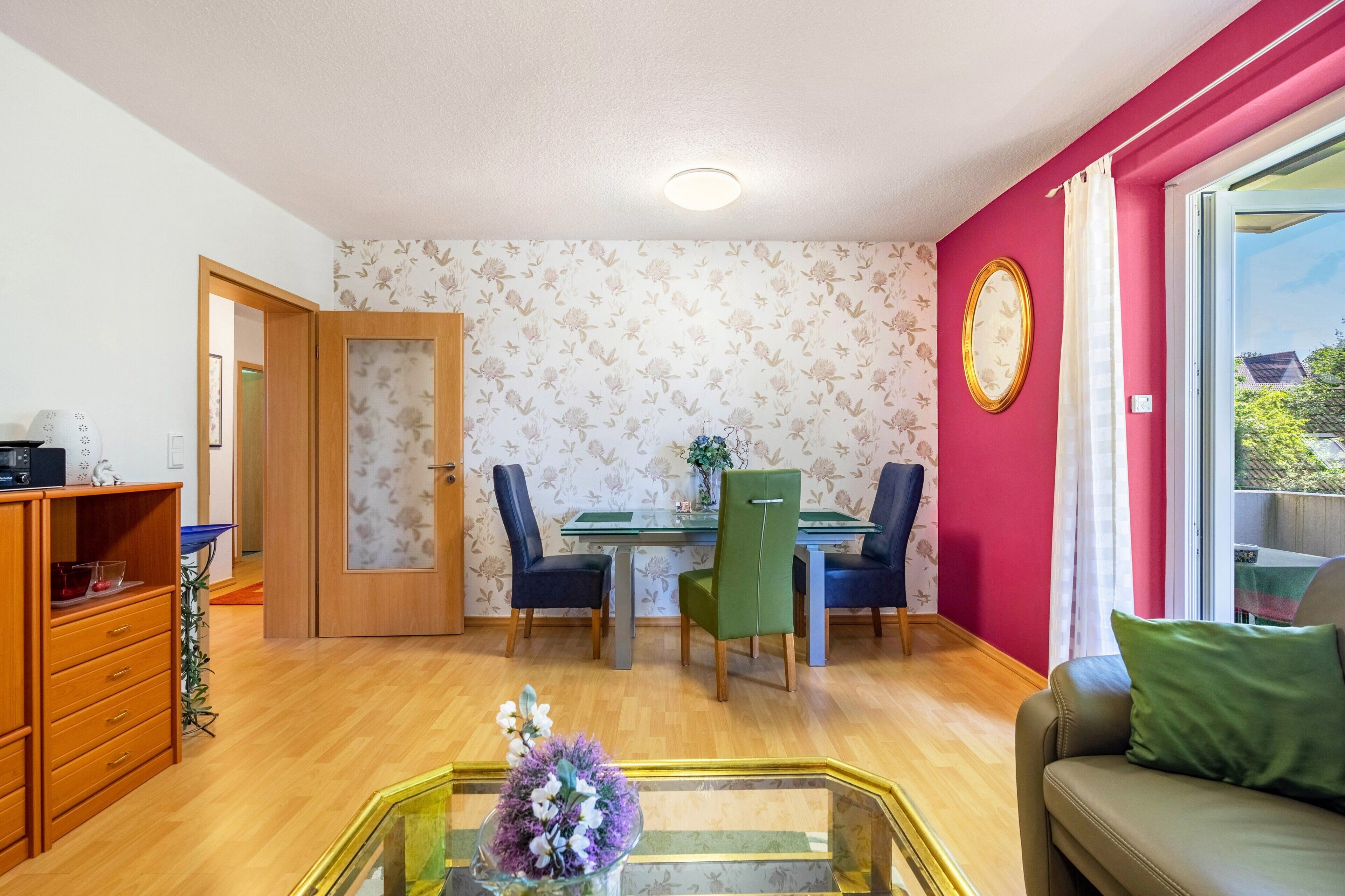 Ein Zimmer mit Esstisch, zwei Stühlen, einer Couch, einem Glastisch mit Blumen, einem Holzschrank und einer Tür zu einem weiteren Raum.