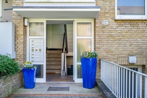 Une entrée d’immeuble avec un escalier, une plaque portant le numéro « 117 », et deux grands bacs bleus ornés de fleurs.