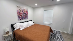 Une chambre à coucher comprenant un lit, une table de chevet, une lampe, une fenêtre avec des stores et une œuvre d’art à motifs floraux accrochée au mur.