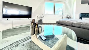 A modern hotel room with a flat-screen TV, a glass coffee table with a remote, a bed, and a large window.