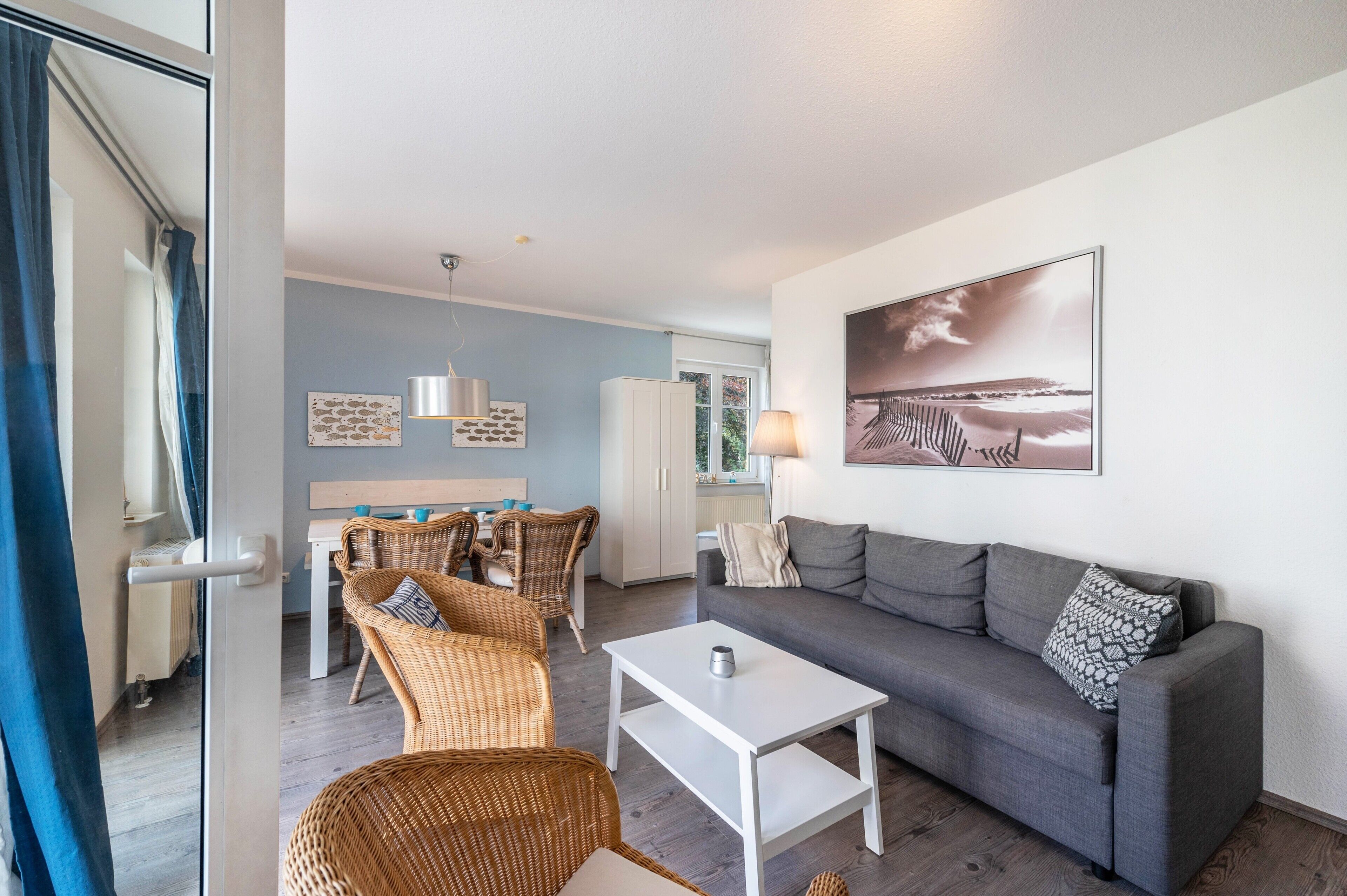 Una sala de estar moderna con un sofá gris, sillas de mimbre, una mesa de centro blanca y un comedor con mesa y sillas.