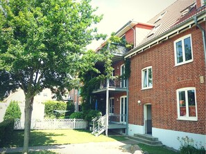 Un bâtiment en briques de deux étages, avec des ornements blancs, un porche à l’avant et un arbre dans le jardin.