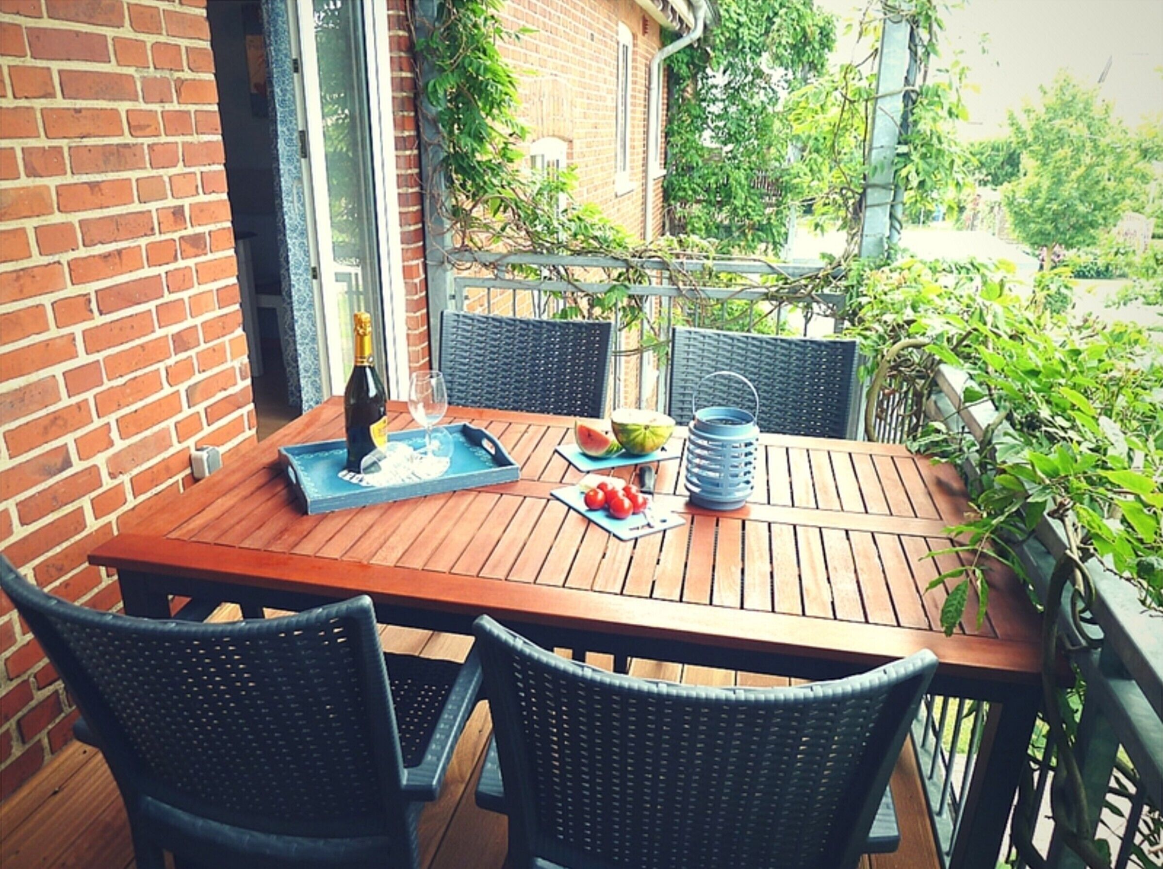 Ein Holztisch mit Stühlen, einer Weinflasche, einem Glas und einer Wassermelone auf einem Balkon mit Ziegelwänden und Pflanzen.