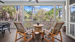Un espace extérieur abrité, aménagé avec des meubles en osier, un ventilateur de plafond et de grandes fenêtres offrant une vue sur les arbres et une maison.
