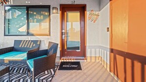 Een overdekt terras met rieten meubels, een deur met welkomstmat en een bord met het nummer 12 erboven.