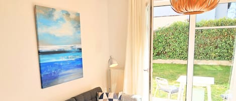 A living room with a sofa, a table with a lamp, a painting, and a view of a garden through a sliding door.