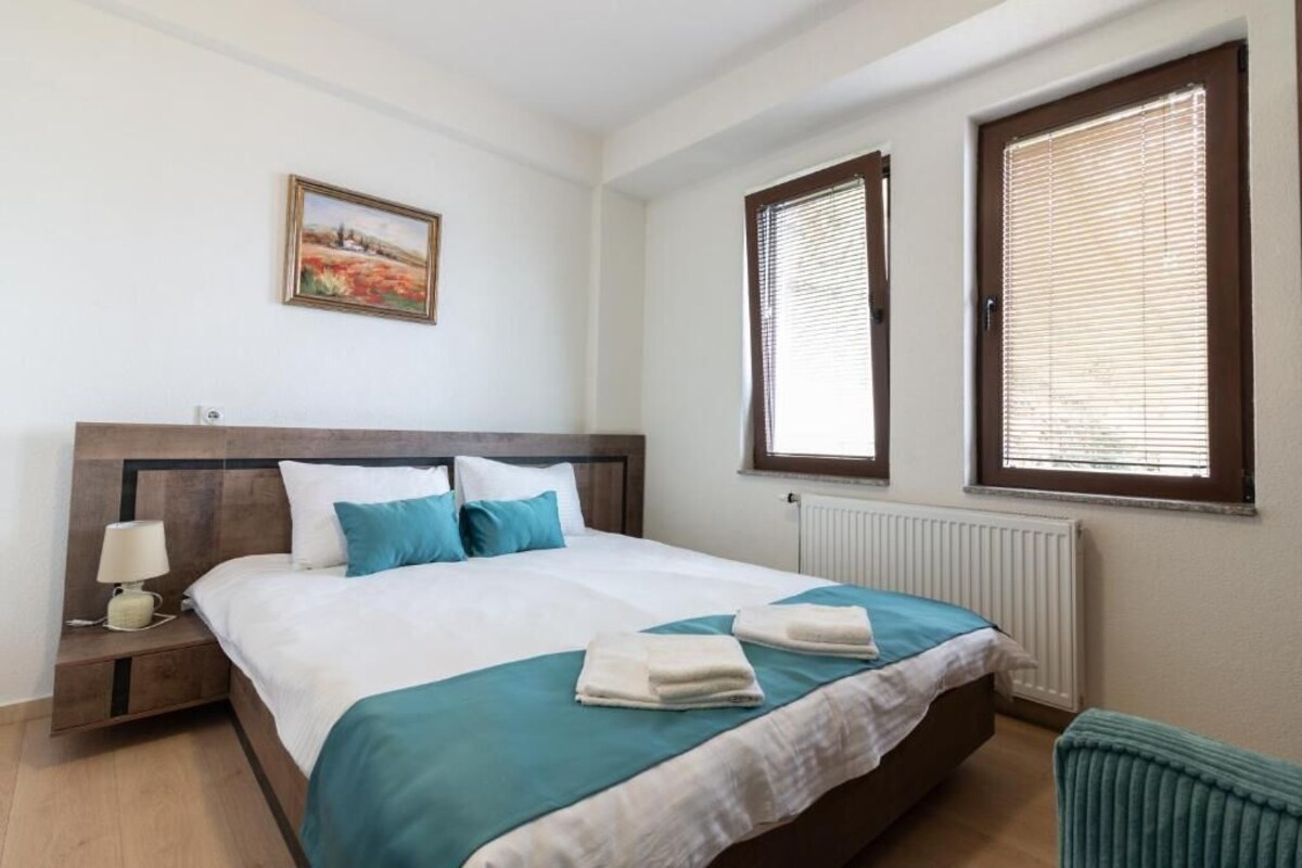 A bedroom with a bed, a nightstand, a lamp, a picture on the wall, and two windows with blinds.