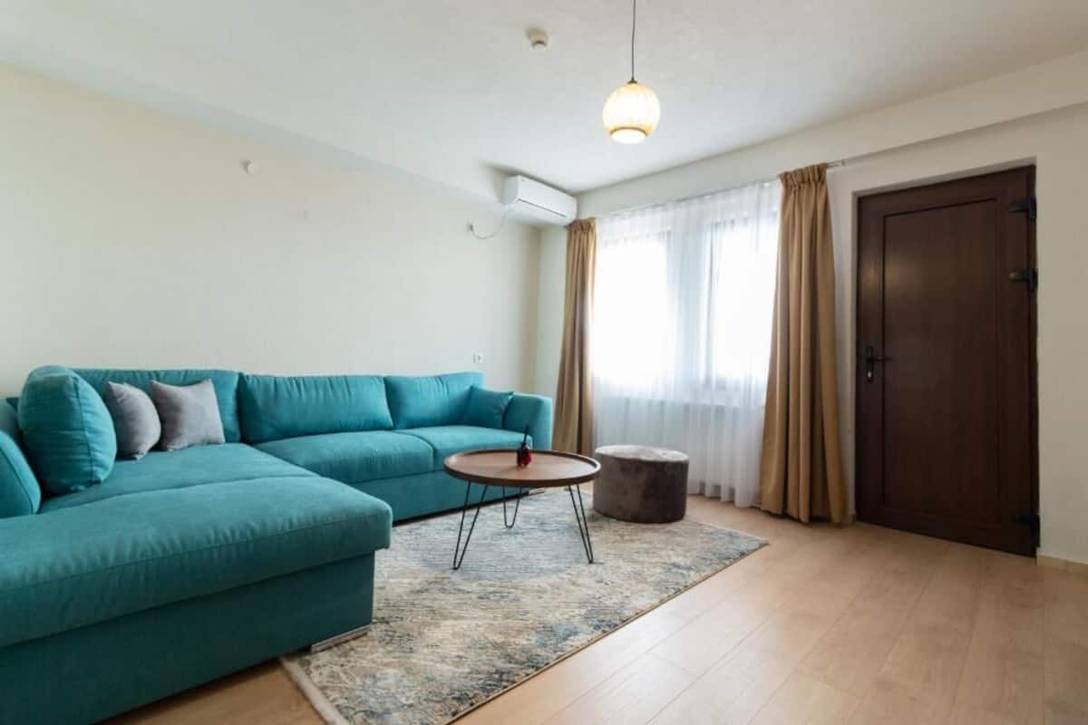 Una sala de estar con un sofá color esmeralda, una mesita redonda, un otoman redondo, un candelabro colgante, una puerta y una ventana con cortinas.