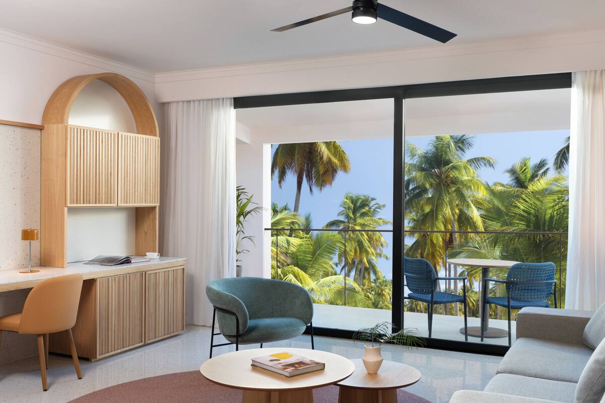 Una habitación moderna con escritorio, sillas, sofá y un balcón con vistas a palmeras, además de un ventilador de techo.