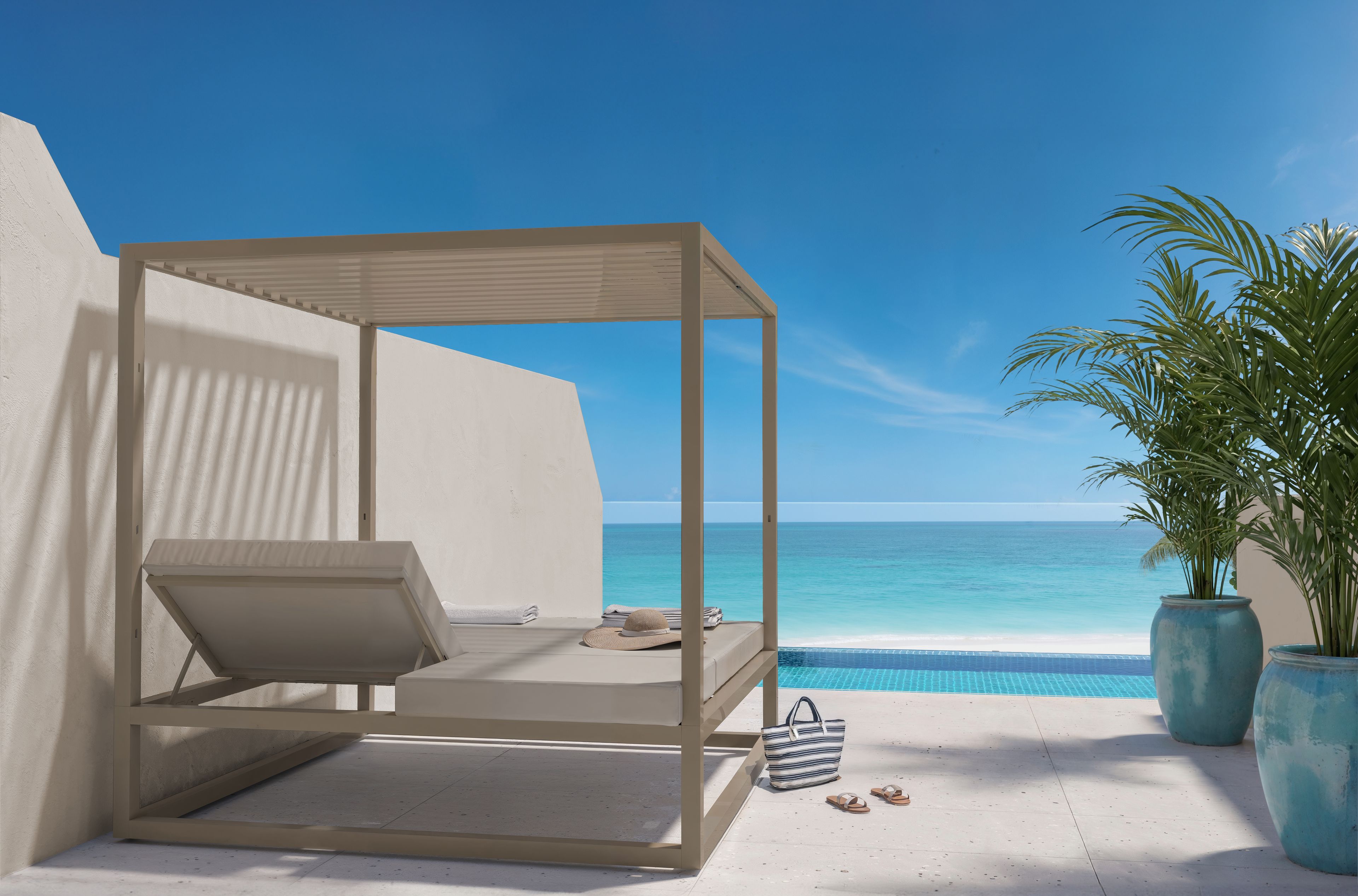 Un balcon doté d’un auvent blanc, une petite table, un chapeau et un sac, d’où l’on peut admirer une mer d’un bleu limpide et une piscine.