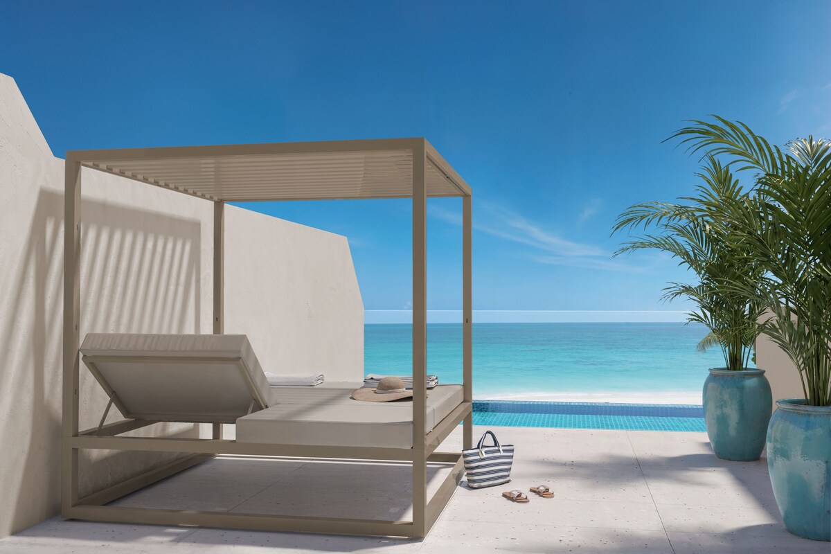 A balcony with a white canopy, a small table, a hat, and a bag, overlooking a clear blue sea and a swimming pool.