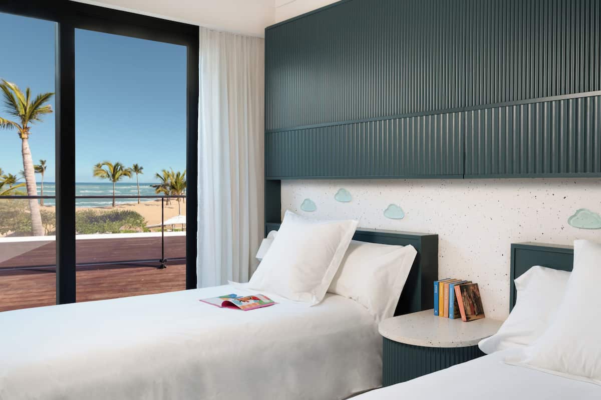 Habitación de hotel con un ventanal grande que ofrece vista a la playa, una cama con ropa blanca y una mesita de noche pequeña con libros.