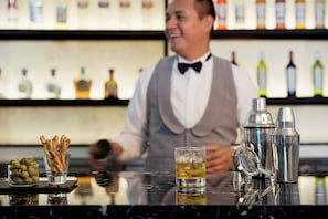 Un coin bar avec un barman, un verre de whisky, des olives et diverses bouteilles en arrière-plan.