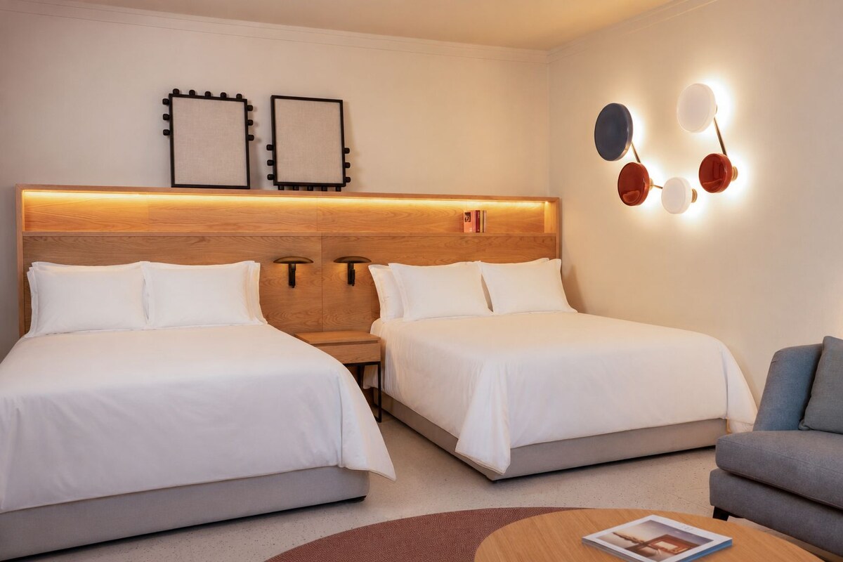 A hotel room with two beds, a wooden headboard, wall-mounted light fixtures, and a small table with a magazine.