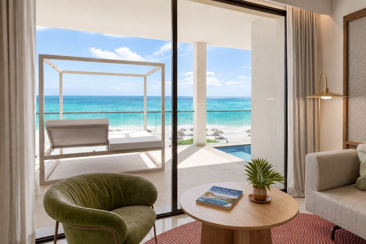 A room with a view of the ocean, a green armchair, a sofa, a round table with a book and a plant, and a sliding glass door leading to a balcony with a pool.
