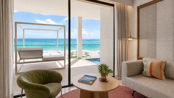 Une pièce avec vue sur l’océan, un fauteuil vert, un canapé, une table ronde avec un livre et une plante, et une porte-fenêtre coulissante donnant sur un balcon avec une piscine.