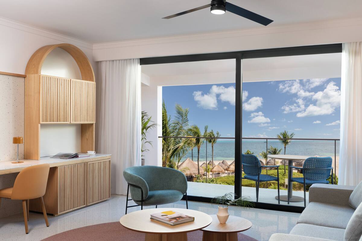 Una habitación moderna con escritorio, sillas, sofá, mesa redonda y balcón con vista a la playa y al océano.