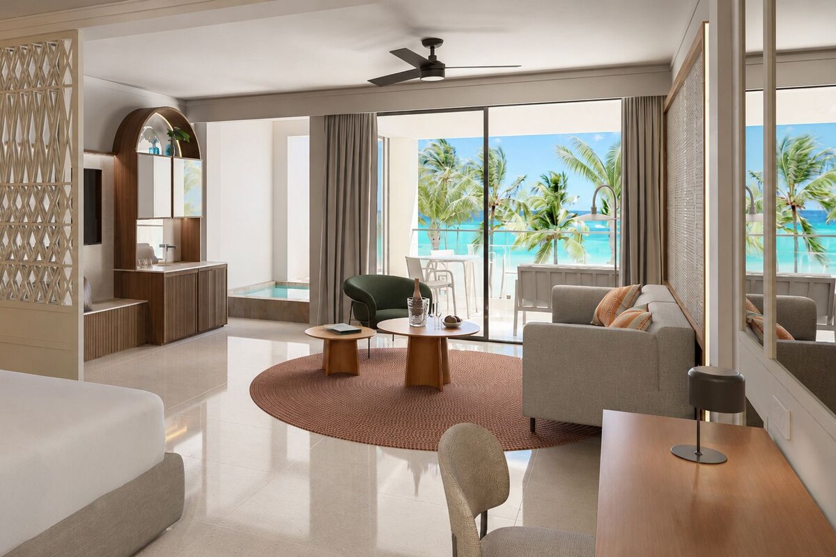 A modern hotel room with a balcony, a bed, a seating area, and a view of the ocean.