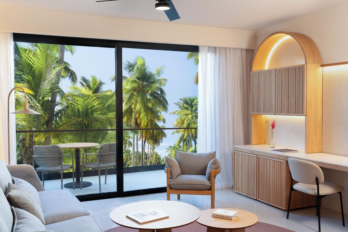 A modern hotel room with a balcony, a sofa, a dining table, and a desk area.