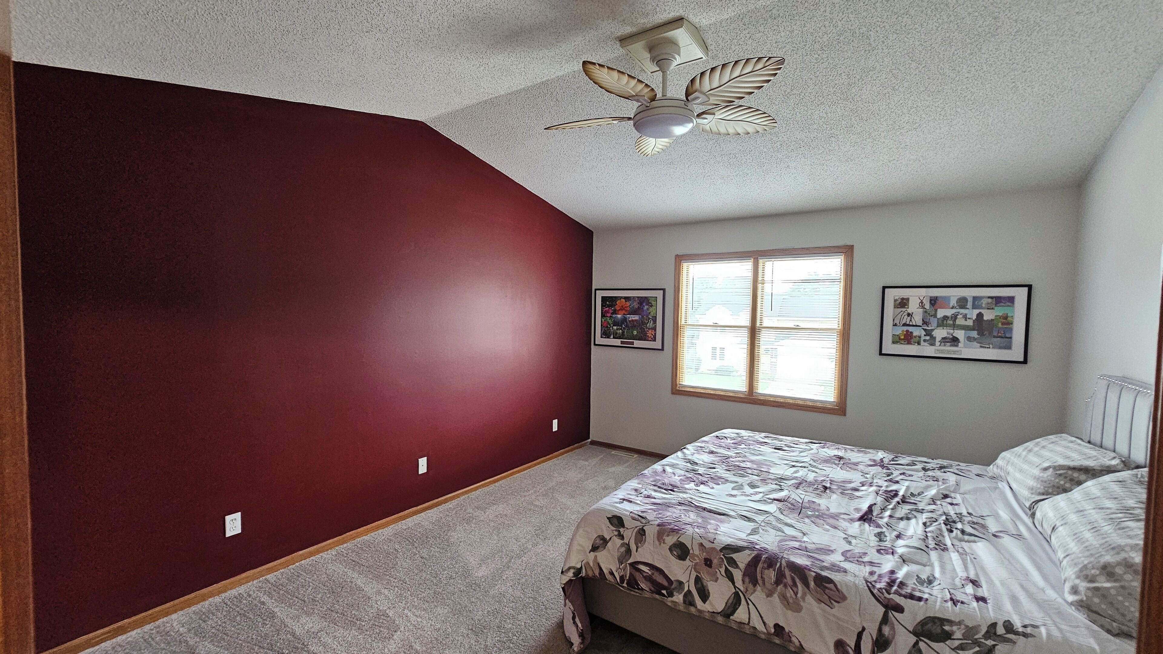 Une chambre à coucher avec un lit, un ventilateur de plafond, une fenêtre avec des stores et deux tableaux encadrés accrochés au mur.