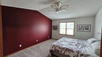 Une chambre à coucher avec un lit, un ventilateur de plafond, une fenêtre avec des stores et deux tableaux encadrés accrochés au mur.