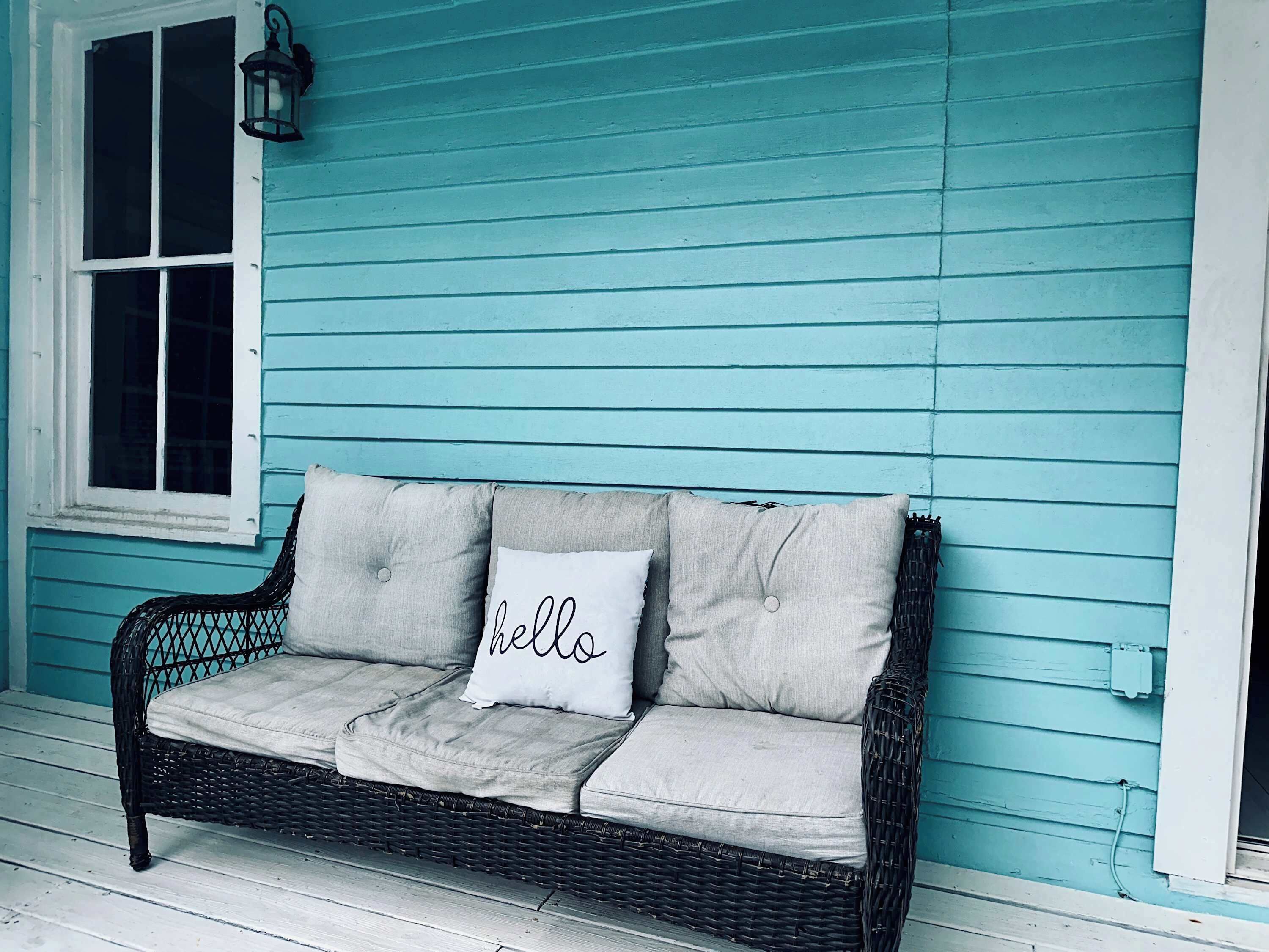 A wicker sofa with a cushion that has 'hello' written on it, positioned against a teal-painted wall with a window to the left.