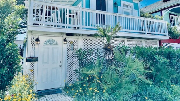 Une maison à deux étages avec un balcon, une porte blanche et un jardin où poussent différentes plantes et fleurs.