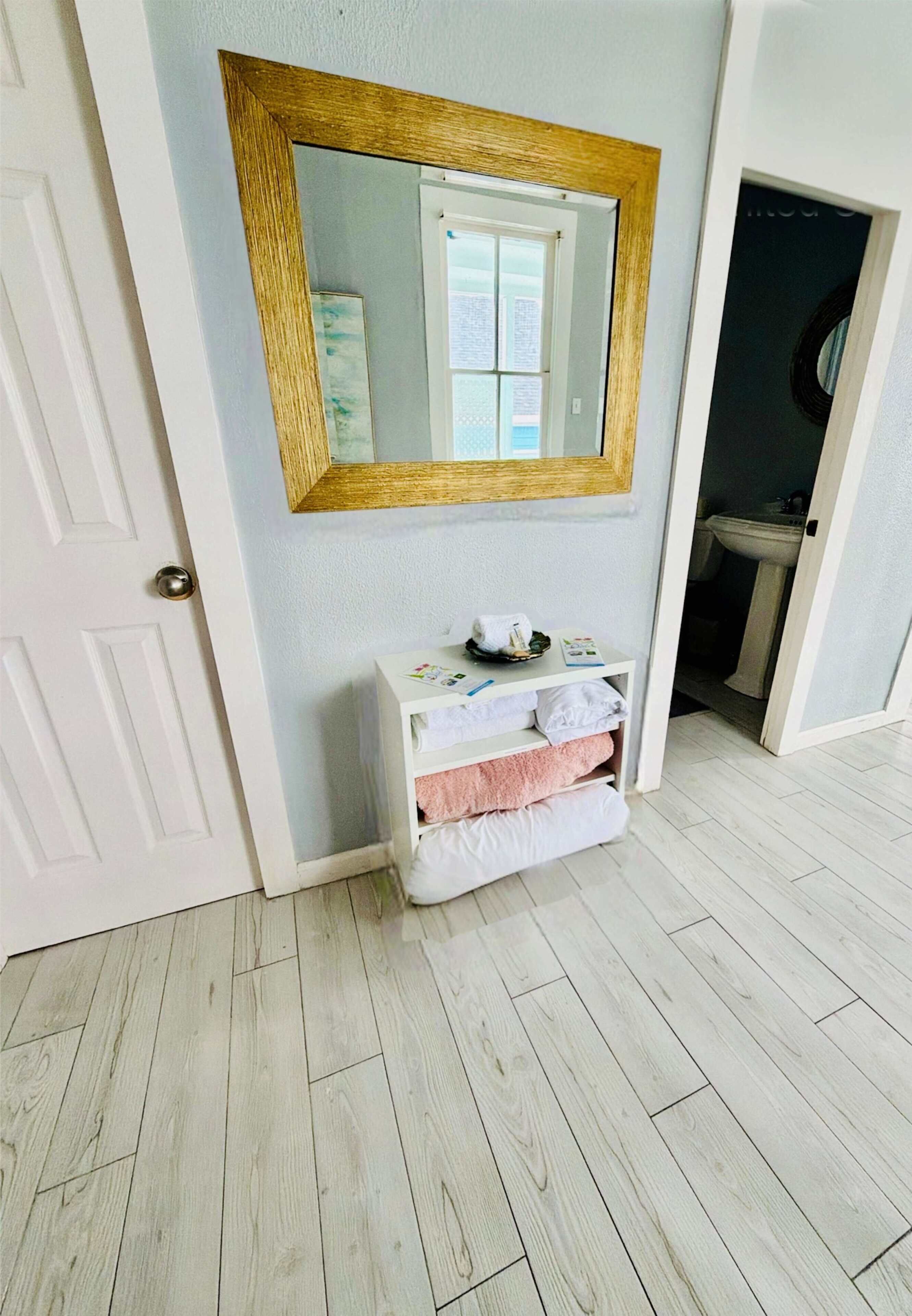 A room with a wooden floor, a white door, a white vanity with towels, a mirror with a gold frame, and a sink in the adjacent bathroom.