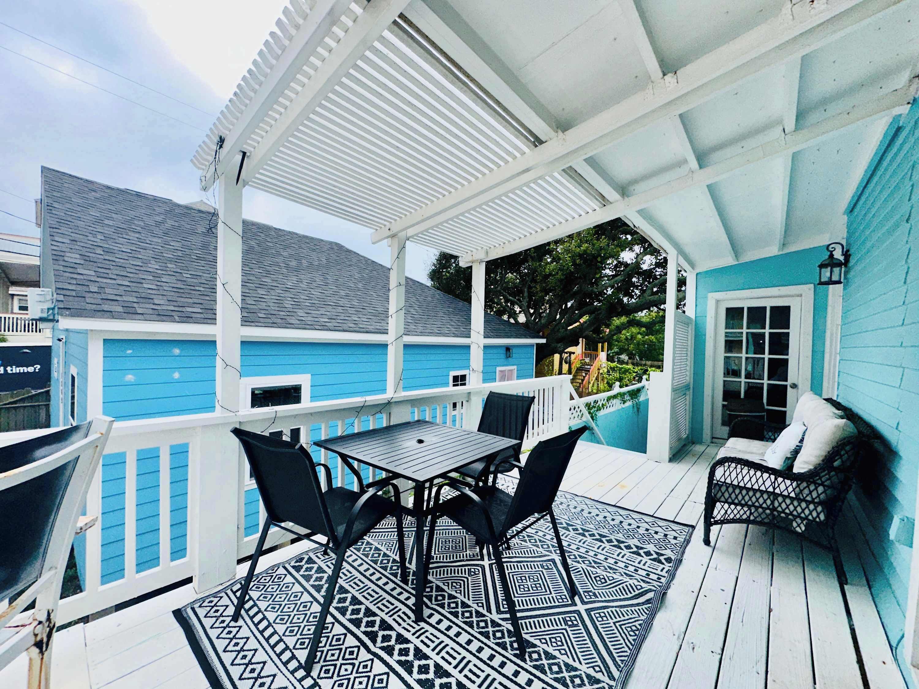 A covered patio with a table and chairs, a patterned rug, and a blue building in the background.
