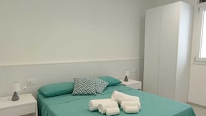 Ein ordentlich gemachtes Bett mit einer türkisfarbenen Tagesdecke, zwei Kissen und aufgewickelten Handtüchern auf einem Nachttisch in Weiß.