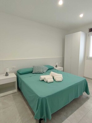 Ein ordentlich gemachtes Bett mit einer türkisfarbenen Tagesdecke, zwei Kissen und aufgewickelten Handtüchern auf einem Nachttisch in Weiß.