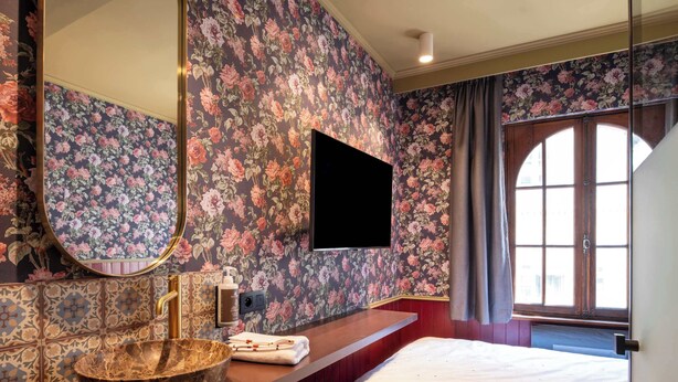 A hotel room with a large bed, a wooden vanity with a sink, a mirror, and a flat-screen TV mounted on the wall. - The Place Brussels (Brussels)