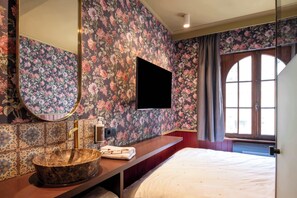 Habitación de hotel con una cama grande, un tocador de madera con lavabo, un espejo y un televisor de pantalla plana montado en la pared.