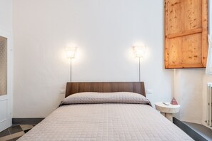 Une chambre à coucher avec un lit, deux lampes fixées au mur, une tête de lit en bois, une petite table ronde avec une lampe et un radiateur.