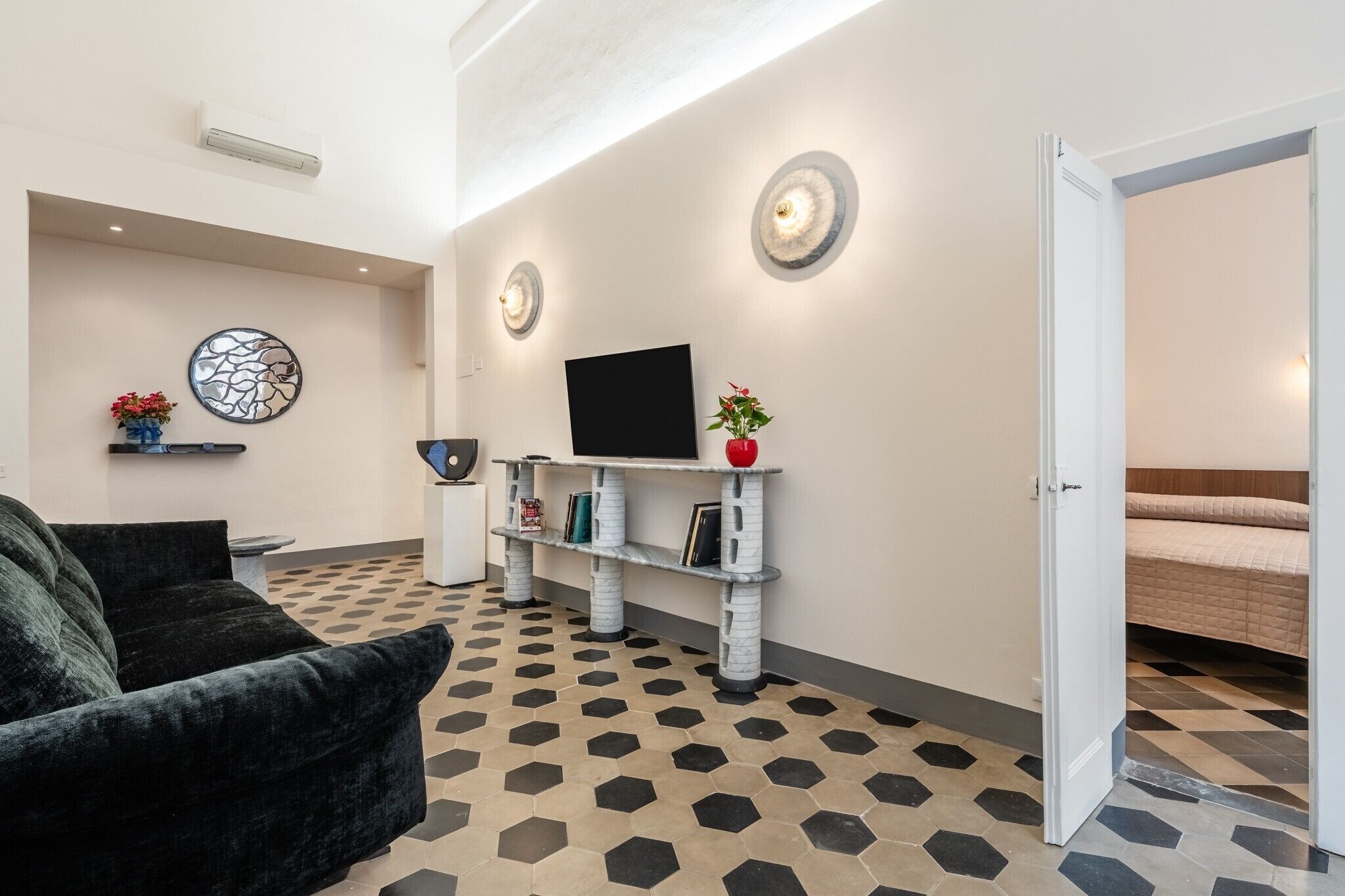 A hotel room with a black sofa, a TV on a white shelf, a bed with a beige duvet, and a patterned floor.