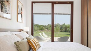Een slaapkamer met een groot bed, witte beddengoed, uitzicht op een grasveld door een schuifdeur en een witte stoel ernaast.