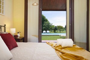 Ein Hotelzimmer mit Bett, Nachttisch mit Lampe und Blick auf ein Schwimmbad und Grünfläche.