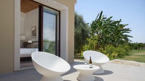 Een moderne kamer met een bed, een glazen deur naar een balkon en twee witte stoelen met een fles en glas op een bijzettafeltje.