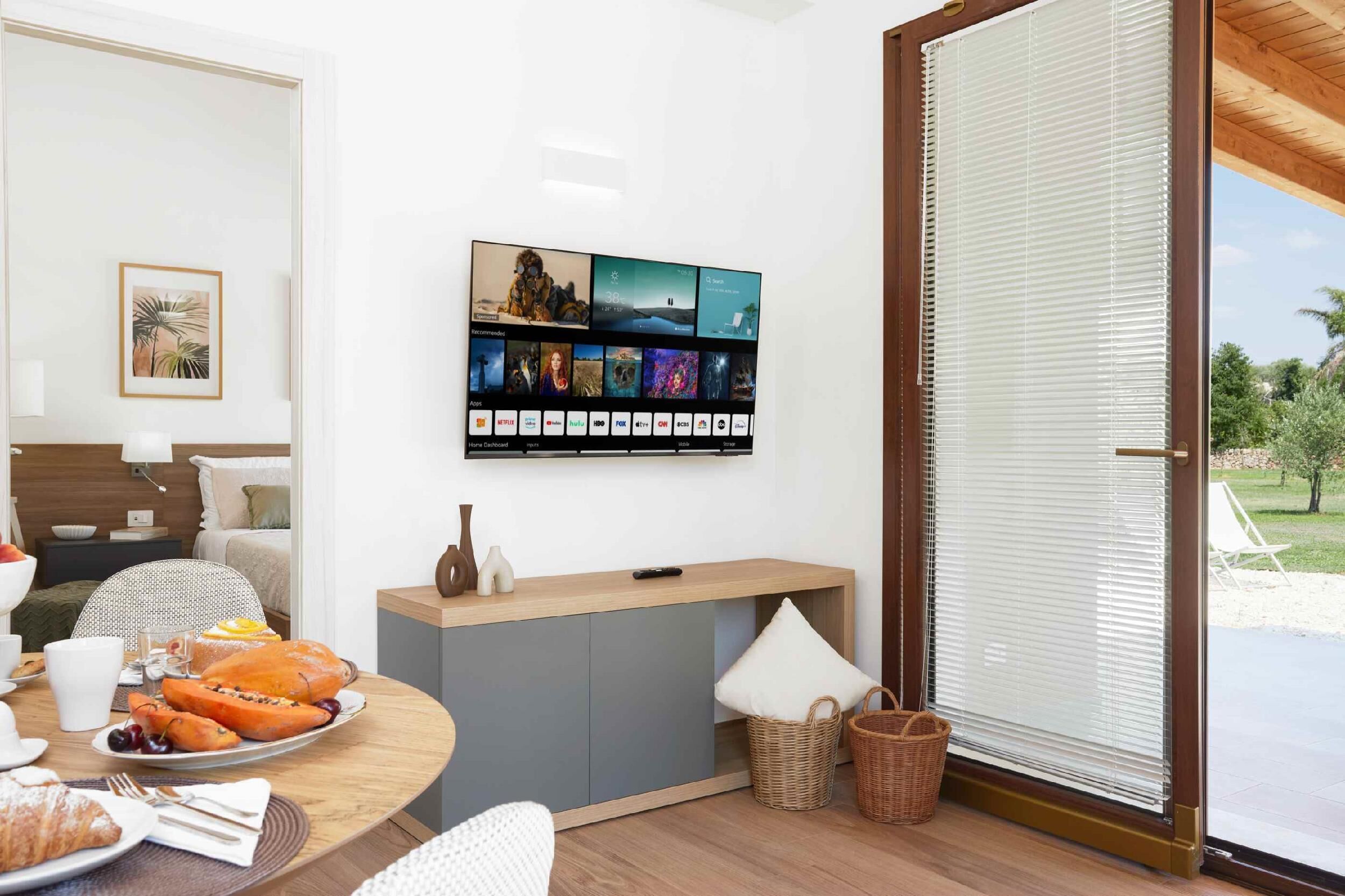 Ein modernes Zimmer mit einem an der Wand befestigten Flachbildfernseher, der verschiedene Sender zeigt, einem Holztisch mit Früchten und Gebäck, einer gemütlichen Sitzecke mit Sofa und Sesseln sowie einem großen Fenster mit Jalousien.