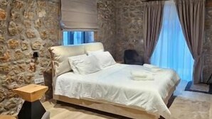 A bedroom with a stone wall, wooden ceiling beams, a bed, a chair, and a window with curtains. - Miltilon (Xylokastro-Evrostina)