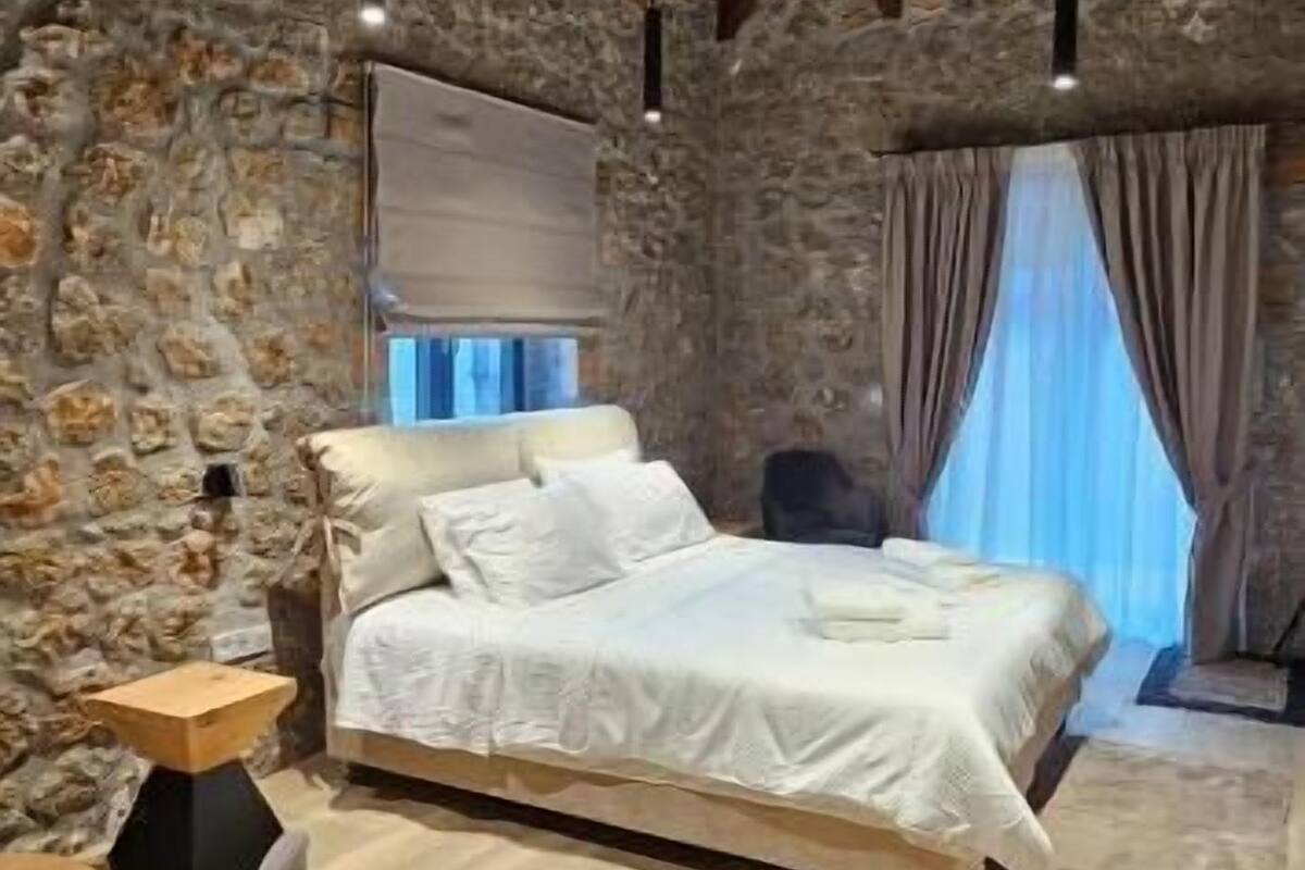 Un dormitorio con pared de piedra, vigas de madera en el techo, una cama, una silla y una ventana con cortinas.