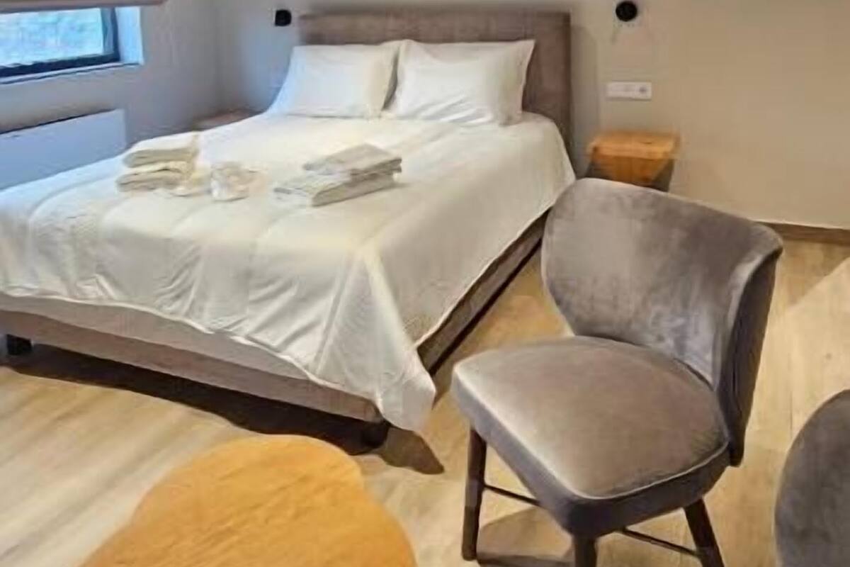Un dormitorio con cama, dos sillas, una mesita redonda, piso de madera y techo inclinado con vigas a la vista.