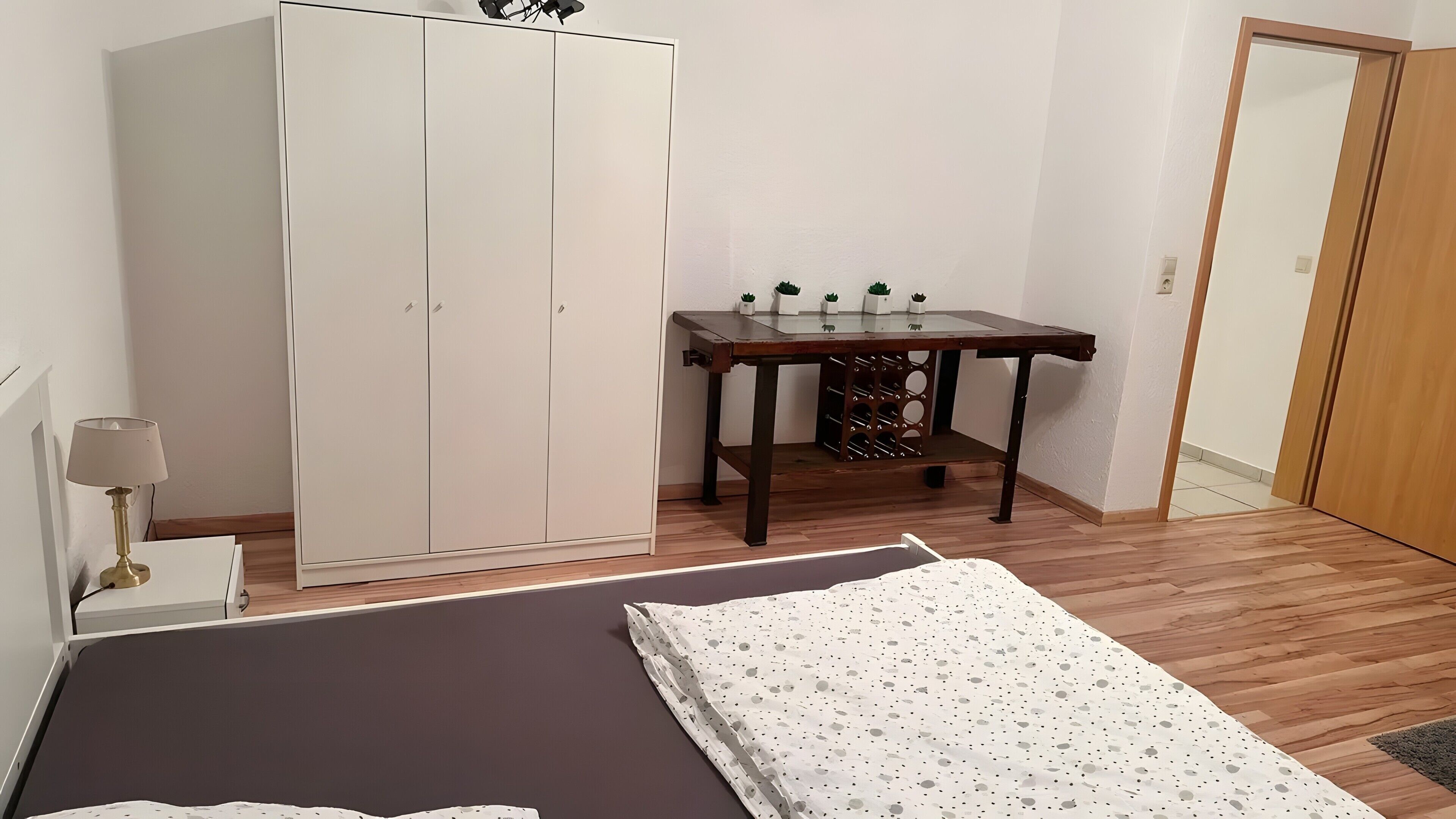 A bedroom with a bed, a wardrobe, a table with potted plants, and a lamp.