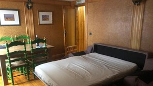 A room with a bed, a desk with chairs, a door, and framed pictures on the wall.