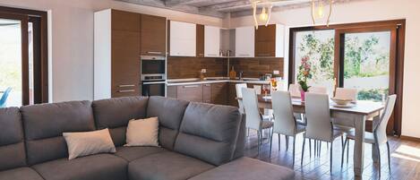 A modern kitchen with wooden cabinets, a dining area with a table and chairs, and a large sectional sofa.