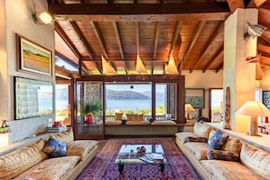 Ein gemütliches Wohnzimmer mit großem Fenster, Holzbalkendecke und Blick aufs Meer.