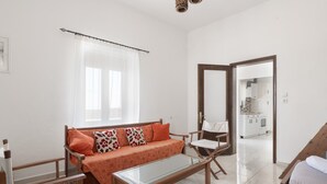A modern, well-lit living room with a sofa, a glass coffee table, and a wooden chair. A kitchen is visible through an open door. - Casa Di Rena Country House: Panoramic Castle View & Endless Blue (Kritinia)