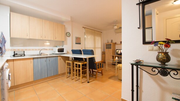 A compact kitchen with a microwave, sink, and wooden cabinets. There is a dining area with a table and chairs. A decorative vase with flowers is on a console table. - Lentiscares 212 by At Home Costa del Sol (Torrox)